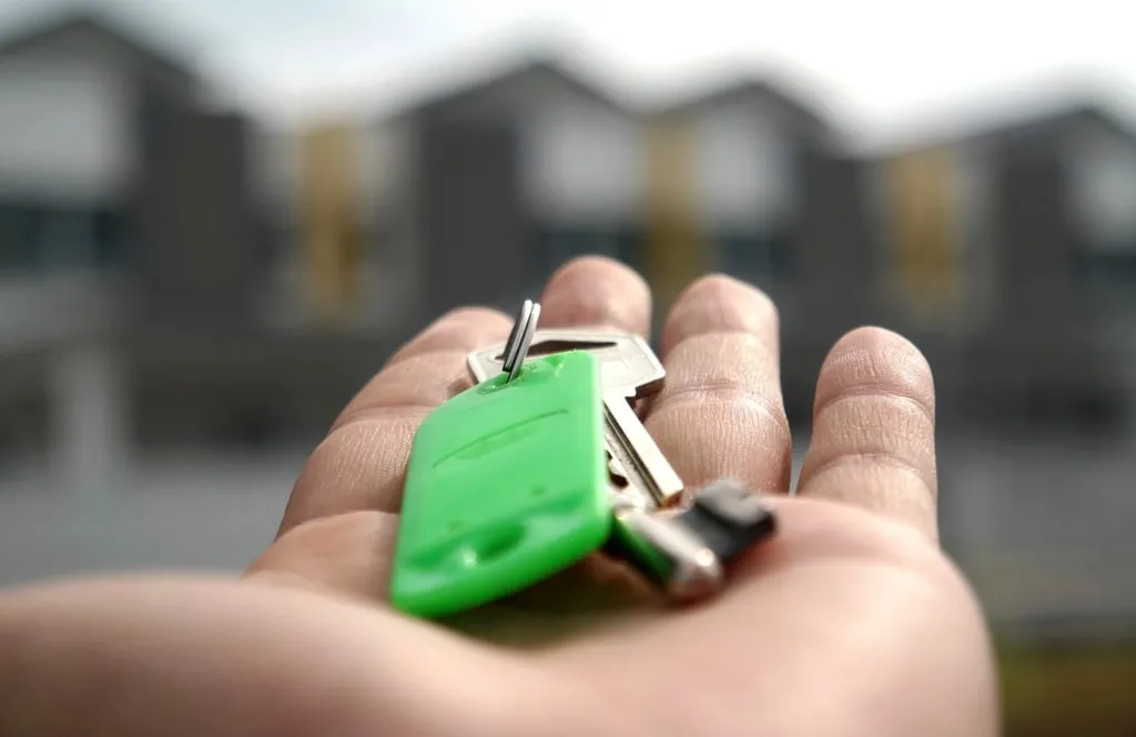 A hand holding a set of house keys on a green key tag, with a row of houses out of focus behind
