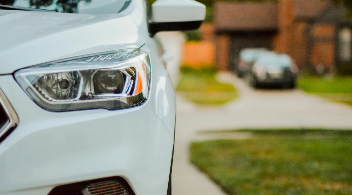 Front of a white car with its headlight in focus, parked on a suburban driveway