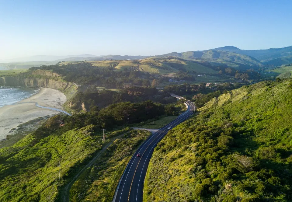 A paved road winding along a green coastal hillside above a sandy bay
