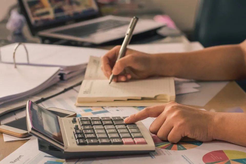Hands using a desk calculator and writing in a notebook beside a binder