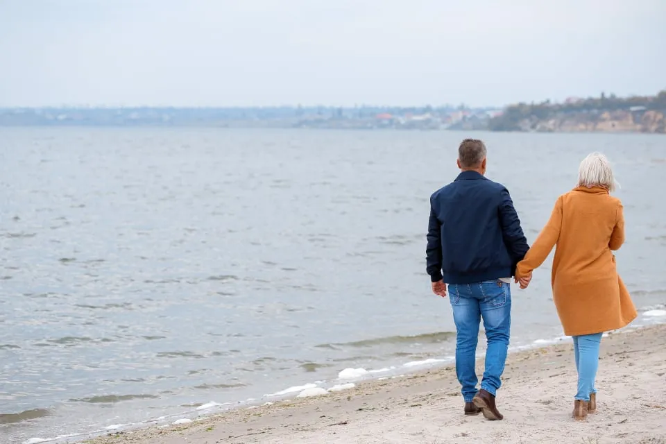 A couple walking hand in hand along a beach at the water's edge, seen from behind, with a town on the far shore