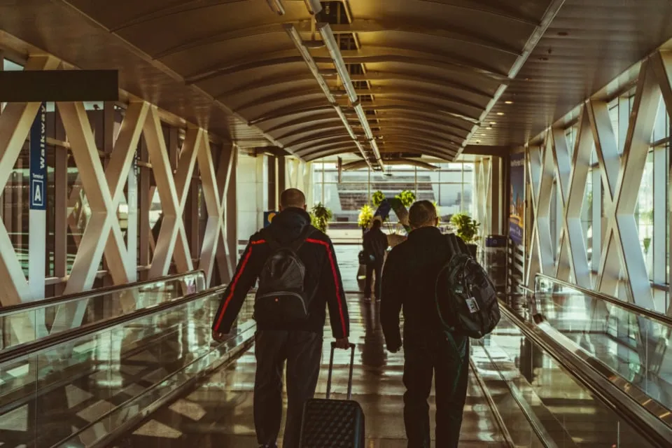 Two travelers with backpacks and a wheeled suitcase walking down an airport moving walkway