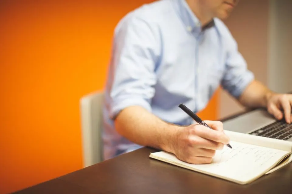 A man in a light blue shirt writing in a notebook next to a laptop