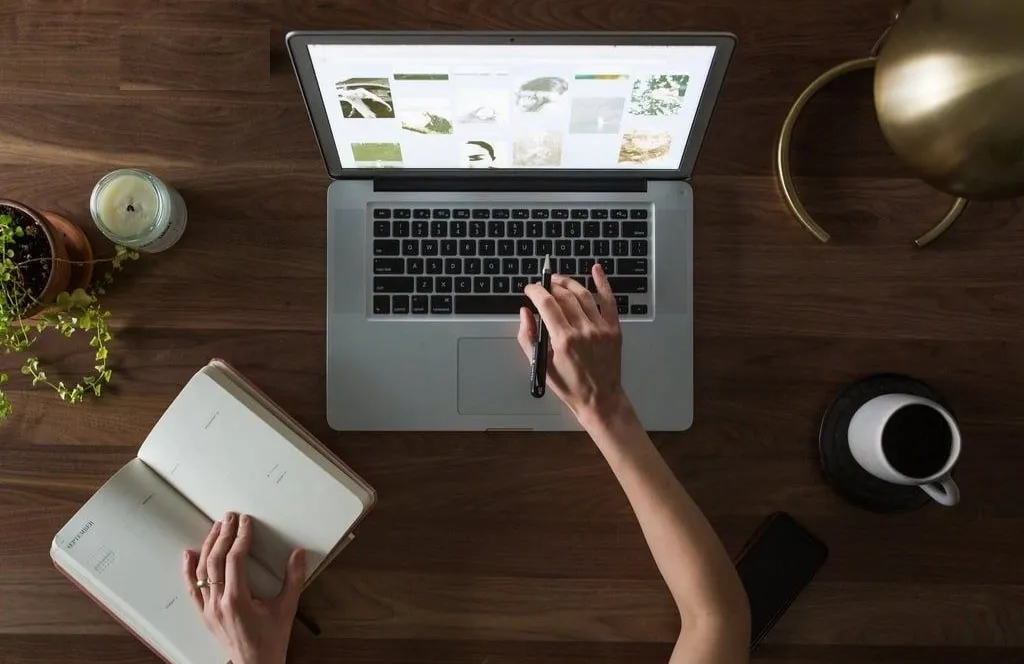 A hand on an open laptop beside a planner and a cup of coffee on a wooden desk