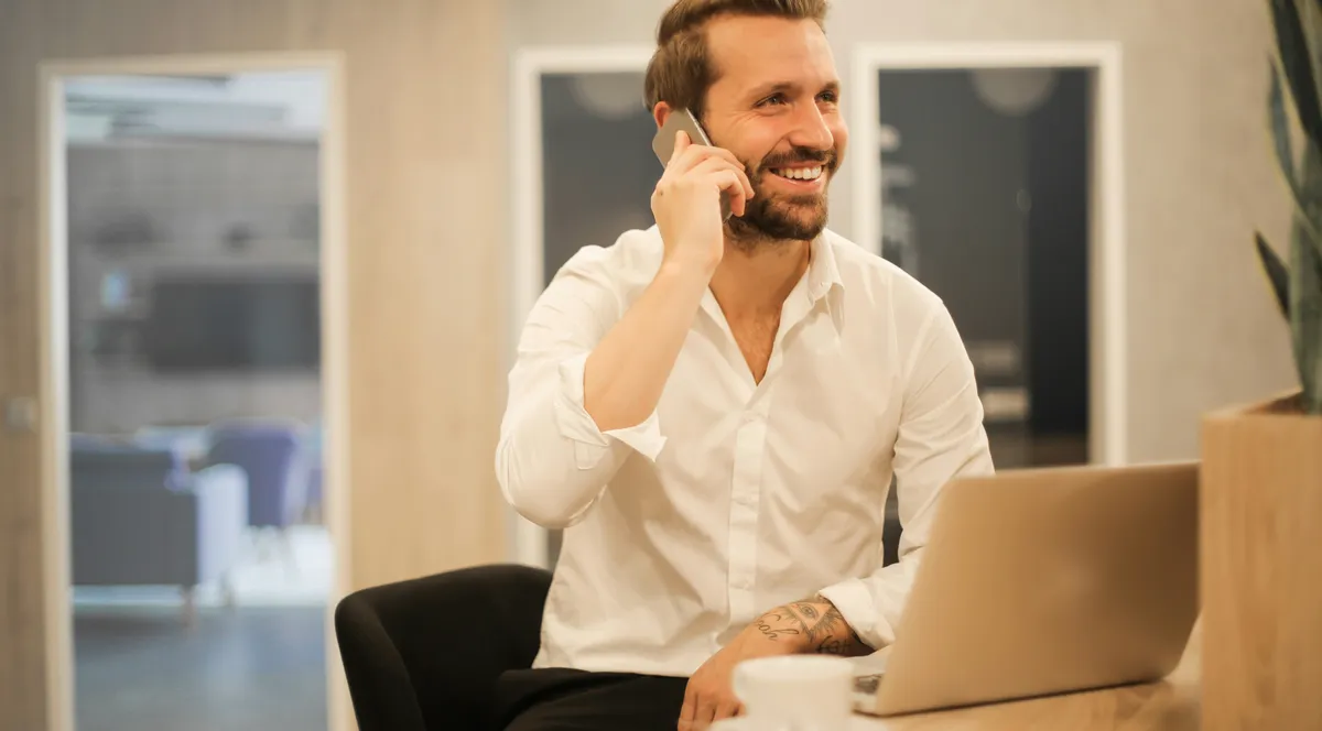 A man in a white shirt on the phone at a desk with a laptop
