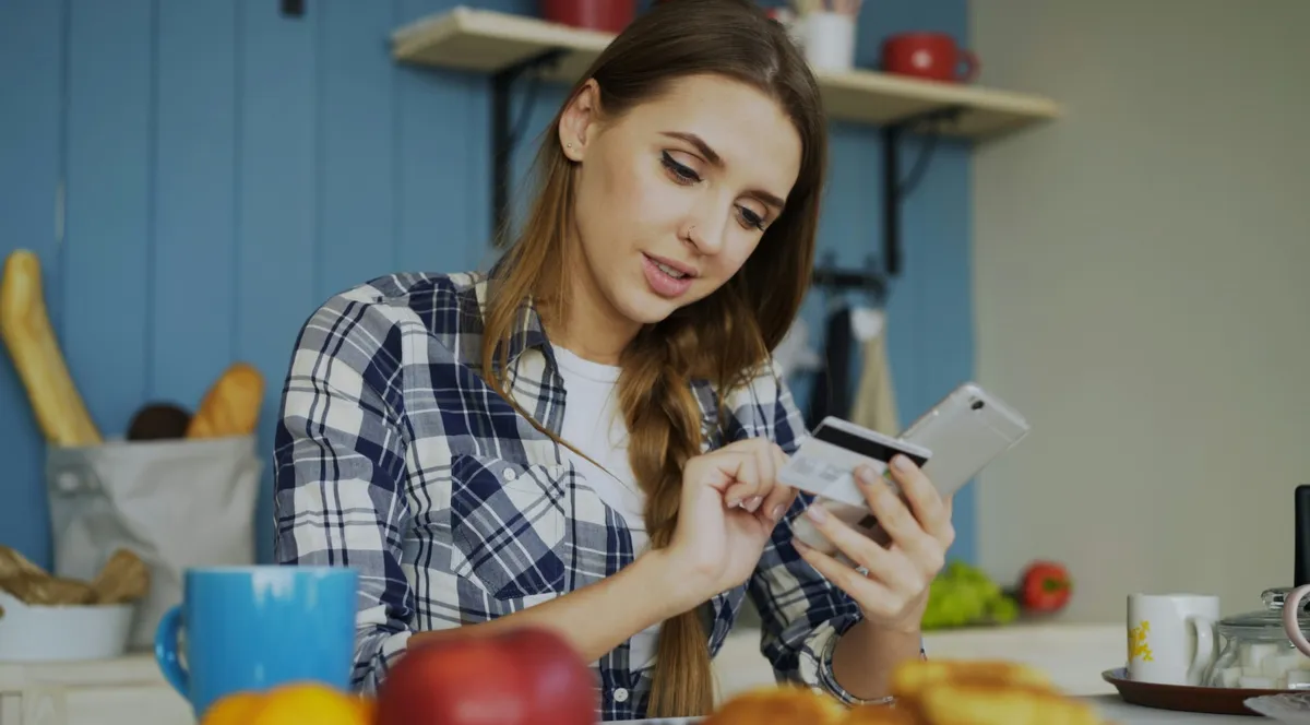A woman at a kitchen table holding a smartphone and a bank card