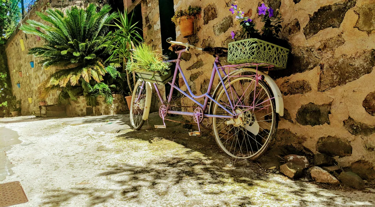 A lilac bicycle with a flower basket leaning against a stone wall in a sunny lane