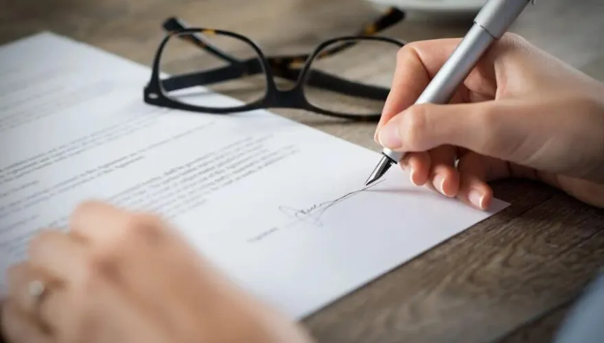 A hand signing a document with a fountain pen beside a pair of glasses