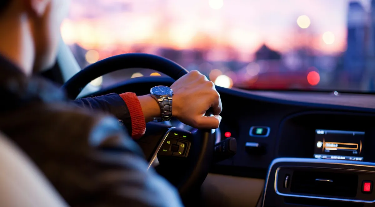 A driver's hand on the steering wheel at dusk, city lights blurred beyond the windshield