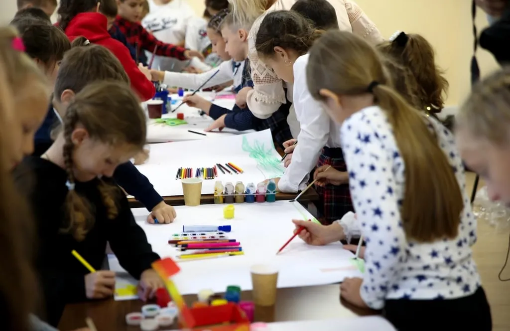 Schoolchildren standing around a long table, drawing and painting on large sheets of paper