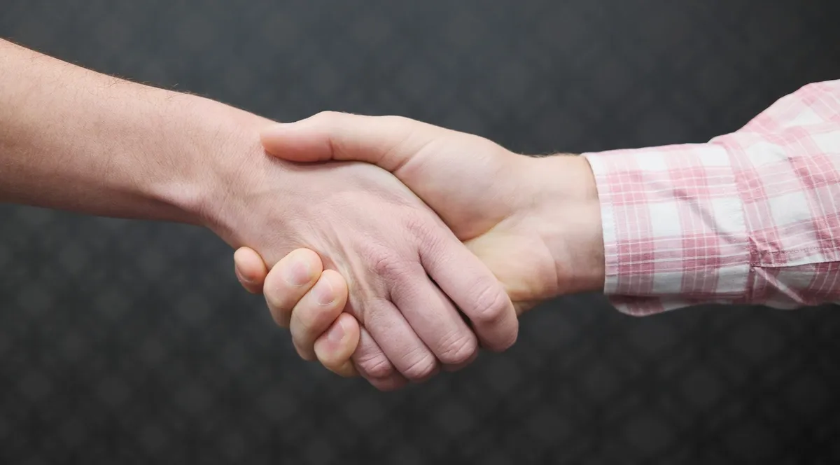 Two people shaking hands against a dark background, one in a pink checked shirt