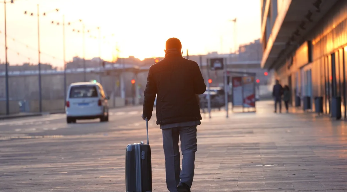 A man seen from behind pulling a wheeled suitcase along a city street at sunrise