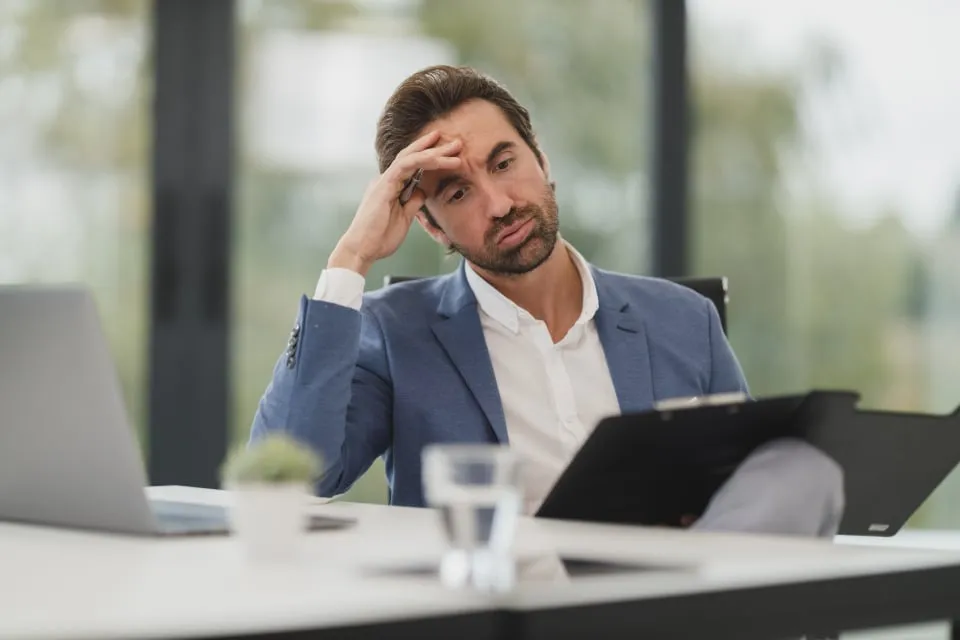 A man in a blue blazer holds his forehead while reading a document on a clipboard at his desk
