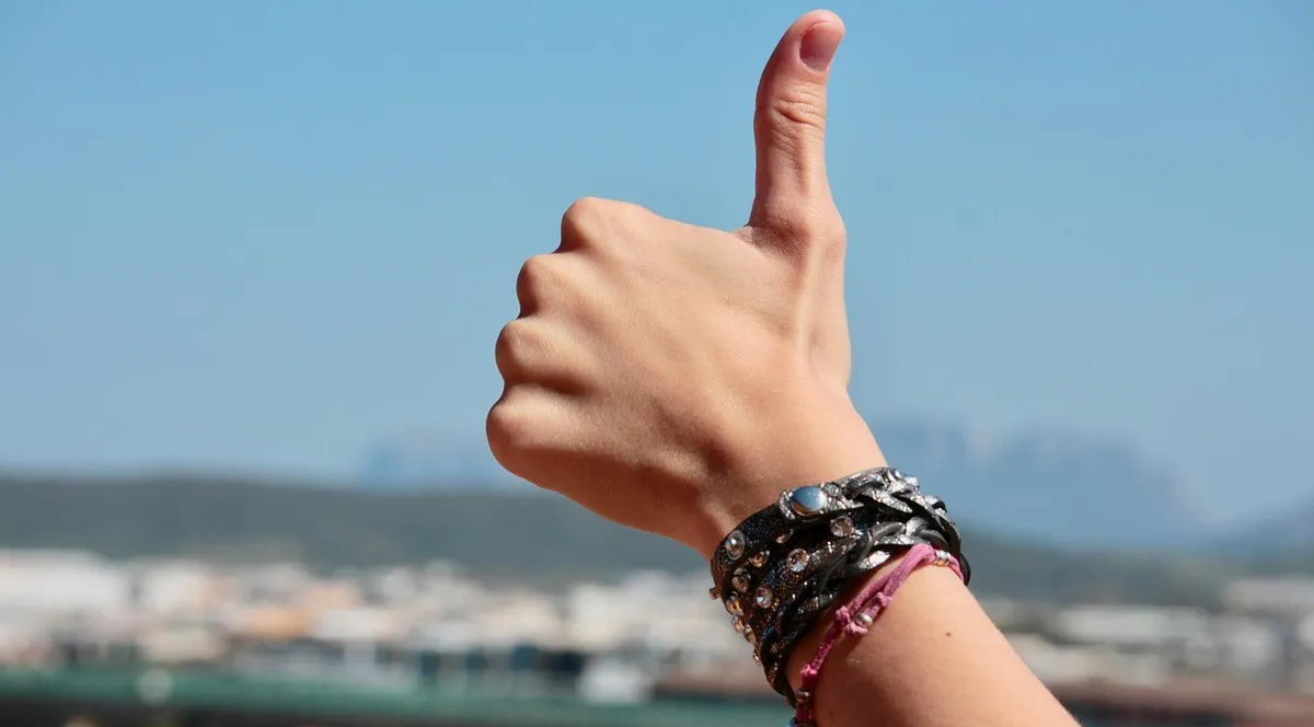 A raised hand giving a thumbs up against a blue sky, with a town and mountains blurred behind
