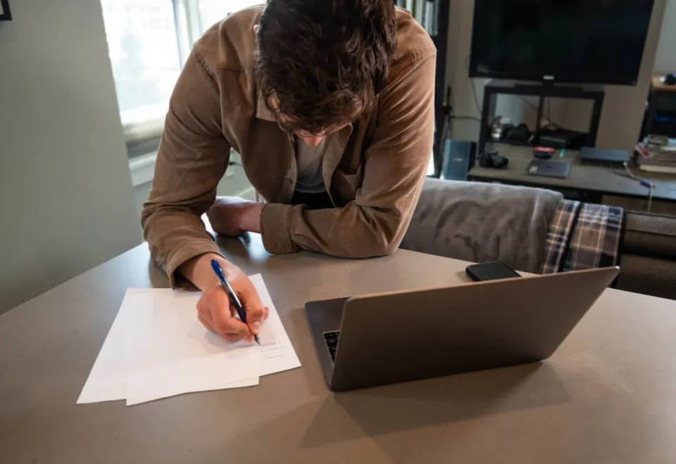 A person leaning over a desk to fill in a paper form beside an open laptop