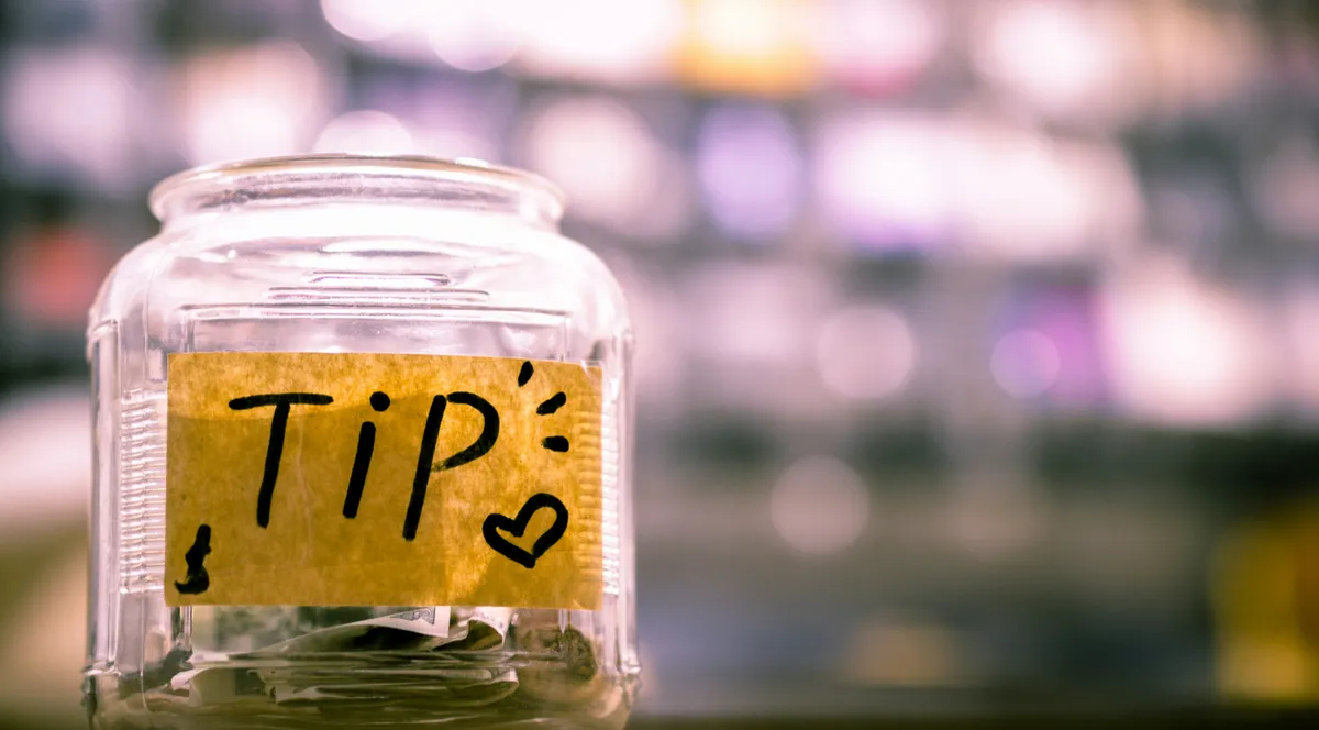 A glass tip jar labeled TIP with a dollar sign, bar lights blurred behind