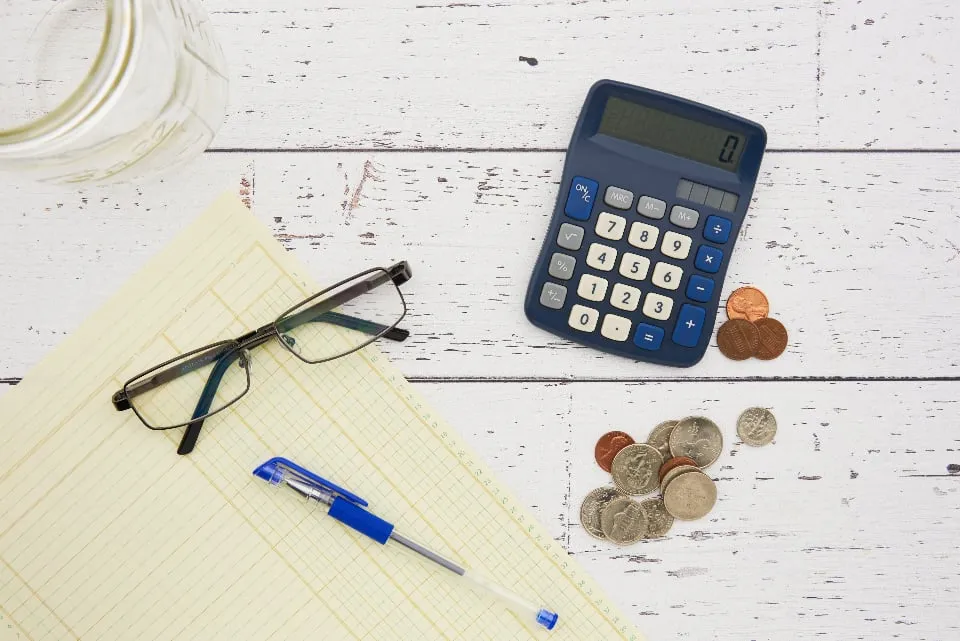 A blue calculator, reading glasses, a pen and U.S. coins on a ledger pad on a white wooden table