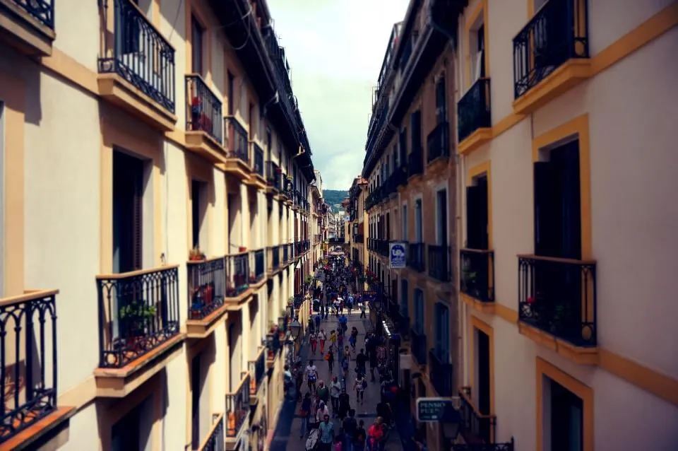 A narrow pedestrian street between apartment buildings with iron balconies, people walking below